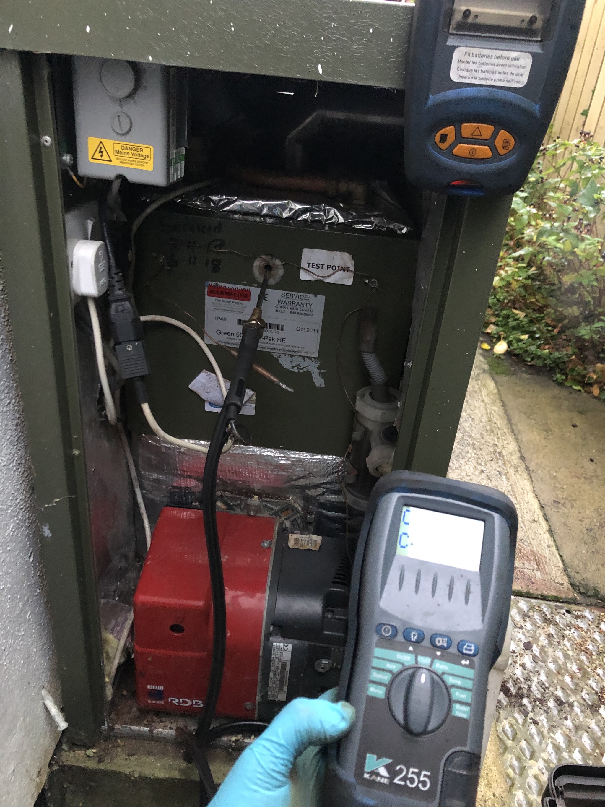 Gloved hand holding a flue gas analyser beside an open external oil boiler during a service