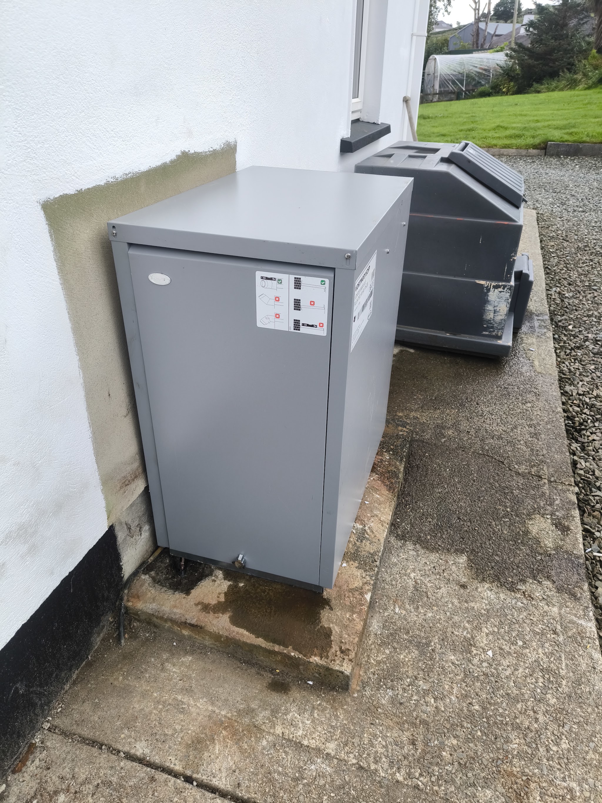 Grey external oil boiler housing on a concrete base against the white wall of a house