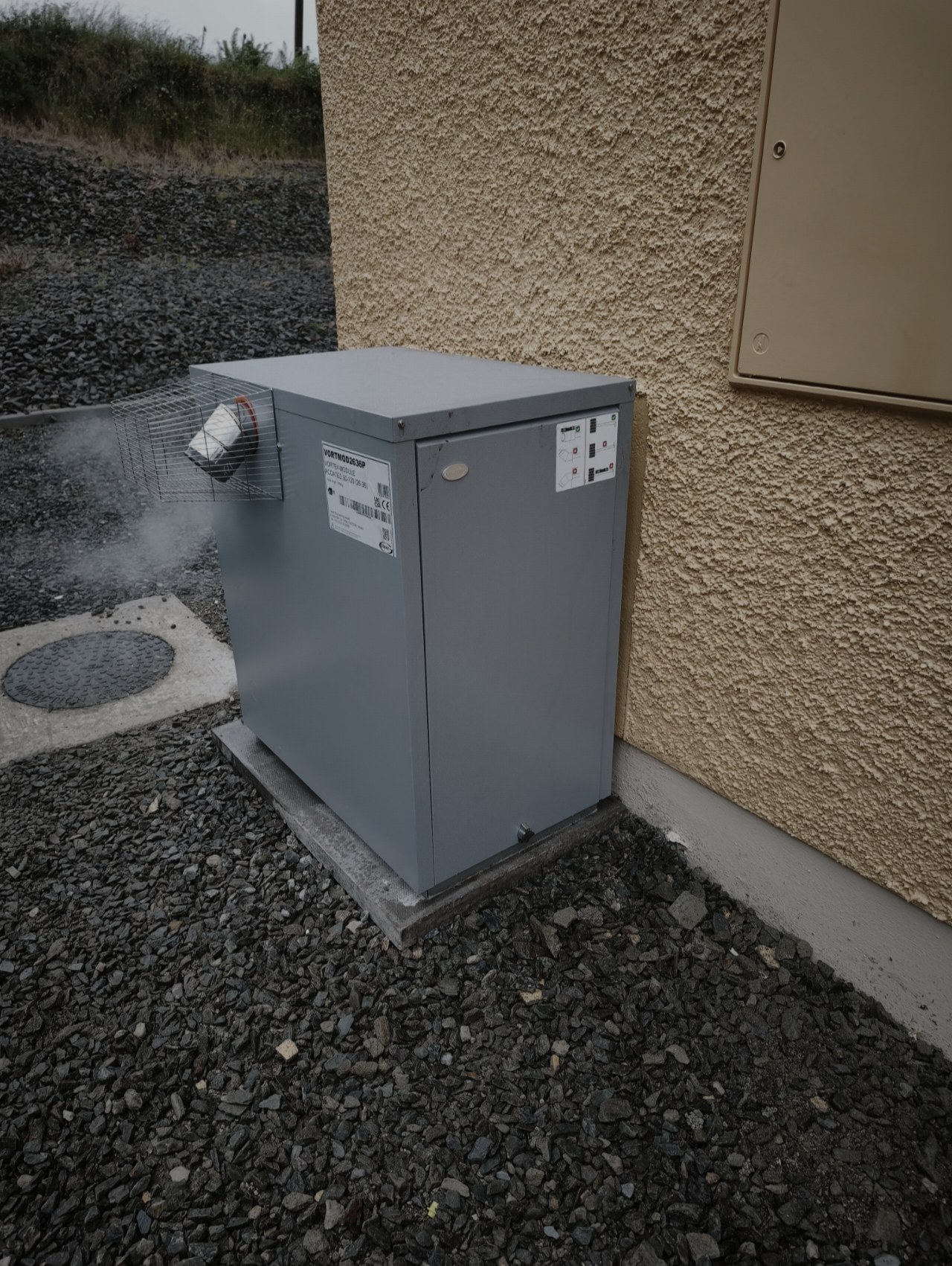 Grey external oil boiler on gravel beside a pebble-dashed wall, with steam rising from the flue